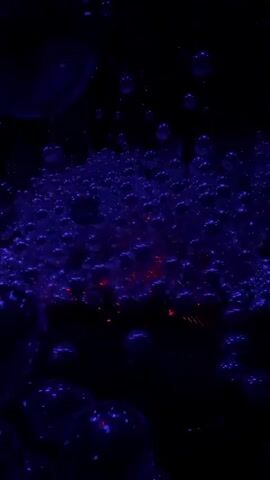 The video displays a close-up, top-down view of a dark, liquid environment filled with numerous small, iridescent bubbles. The bubbles, appearing in shades of deep blue and purple, are illuminated by an unseen light source that creates shimmering reflections. Scattered amongst the bubbles are tiny, glowing red specks, possibly from another substance or a different lighting effect. Some larger, more prominent bubbles are visible, particularly towards the bottom of the frame, catching the light and appearing almost spherical. The overall impression is one of a dynamic, abstract composition of light and texture within a dark setting.