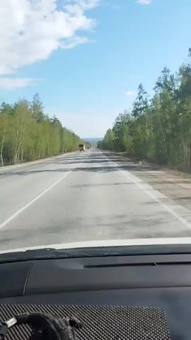 The view from inside a car traveling on a straight two-lane highway is depicted in the video.  The road is flanked by trees on both sides. The sky is blue with some white clouds. A yellow dump truck is initially seen in the distance, followed by a dark vehicle. The dashboard of the car, including a black-and-gray patterned object, is visible at the bottom of the frame.