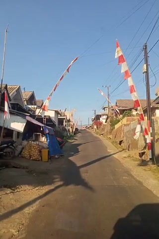 The video is a first-person perspective of someone traveling down a narrow, paved road in a village. The road is flanked by houses and vegetation. Several red and white Indonesian flags are visible, hung across the road and on poles, suggesting a national holiday or celebration. The sky is clear and blue. The camera angle is tilted, making the scene appear vertical. Objects like a blue tarp, bags of produce, and a motorcycle are visible near the side of the road. The sun is casting shadows, indicating it is likely morning or late afternoon.