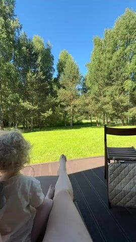 A person with blonde, curly hair and a white shirt sits on a wooden deck next to another person's leg with bare feet, looking out at a grassy yard with trees under a blue sky. A chair is visible on the right. The video then zooms in on the grassy yard and trees, with a small red object partially visible hanging from a tree in the middle of the frame.