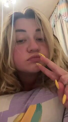 This media file features a close-up shot of a woman with blonde hair and rosy cheeks. She is making a peace sign with her fingers and has bright yellow painted nails. She is wearing a purple top. The camera then pans to show her lying down with several clear cups, possibly for cupping therapy, applied to her back. In the background, a wooden floor and furniture can be seen, suggesting an indoor setting, possibly a bedroom. The overall mood appears casual and relaxed. No visible text is present in the media.