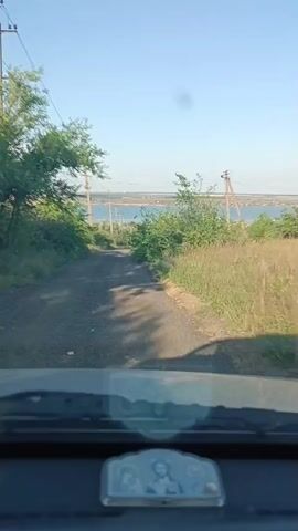 The video shows a point-of-view perspective from inside a vehicle driving down a dirt road. The road is flanked by lush green vegetation and trees on either side. In the distance, a body of water, likely a river or bay, is visible under a clear, blue sky, suggesting a sunny day. Power lines run alongside the road, indicating a rural or semi-rural setting. The overall mood is serene and peaceful, evoking a sense of a journey through the Ukrainian countryside, specifically Bohnatove, Ukraine. The time of day appears to be late afternoon or early evening, judging by the soft lighting and shadows. No people are visible in the video, but the presence of a small decorative religious icon on the dashboard suggests a personal touch to the vehicle.