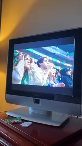 A flat-screen Philips television is positioned on a dark wooden surface. The television displays a soccer match, showing players in green and yellow uniforms on a bright green field. In the foreground of the televised scene, a woman with blonde hair, wearing a white and green striped shirt, is visible with her hands on her head, appearing distressed. Other spectators are also in the background of the televised image. On the surface below the television, there are a few small, unwrapped food items. The room is dimly lit, with a warm yellow light source casting a shadow on the wall behind the television. The television screen is on and actively displaying content.