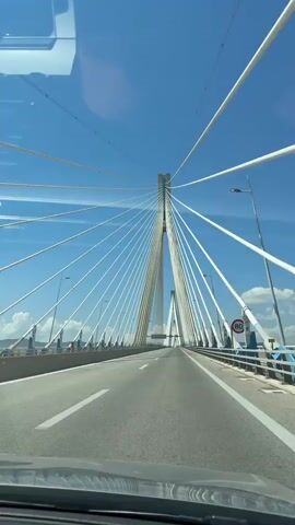 The video is filmed from inside a vehicle driving on a modern cable-stayed bridge. The camera is pointed forward and slightly to the right, capturing the road ahead, the bridge's infrastructure, and the sky. The bridge features tall, white pylons with numerous white cables fanning out to support the roadway. The roadway is asphalt with clear white lane markings. On the right side of the bridge, there is a blue and white railing and a speed limit sign indicating 80. The sky is bright blue with some white clouds visible. The overall impression is a clear, sunny day with a clear view of the expansive bridge structure. The video is short and appears to be a continuous shot without any significant cuts or changes in perspective other than the natural movement of the vehicle.