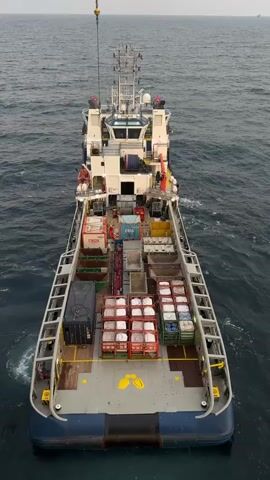 An aerial view shows a large ship with a wide deck loaded with cargo. Two workers in orange jumpsuits and white hard hats are visible on the deck, one near the bow and another further back. The deck is filled with various containers, including several large CBOX containers, some stacked on top of each other. There are also numerous smaller crates, some with white contents, and other industrial-looking equipment. The ship is at sea, with the ocean visible all around. A crane arm extends from above, suggesting cargo is being loaded or unloaded. The overall scene depicts an active maritime operation.