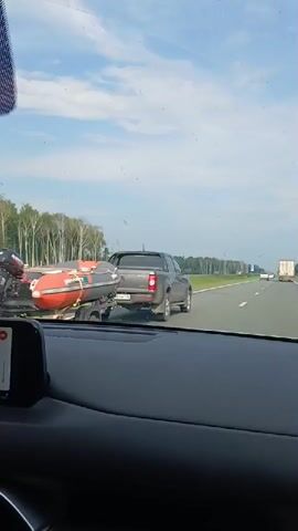 The video shows a view from inside a car driving on a highway. The sky is partly cloudy. On the highway ahead, there are several vehicles, including a white truck. To the left of the highway, there is a grassy area and a line of trees. The car's dashboard is visible in the foreground. The video appears to be filmed during the daytime.