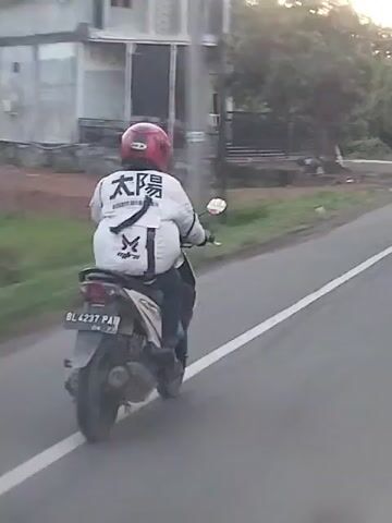A person wearing a red helmet and a white jacket with black Japanese characters "太陽" (Taiyō) and a black symbol is riding a light-colored scooter on a paved road. The scooter has a visible license plate, "BL 4237 PA." The rider is moving away from the camera, which is positioned behind and slightly to the right of the scooter. The road features a solid white line on its right edge. The background consists of green foliage from trees and some distant buildings under warm, direct sunlight, suggesting either morning or late afternoon.