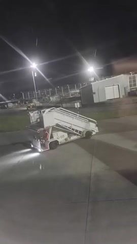 At night, a mobile stairway truck is stationary on a large concrete surface, possibly an airport tarmac. The truck, painted white, has a set of stairs on an incline, extending upwards. The vehicle is illuminated by its own headlights, casting a glow on the concrete. The truck is labeled "42-059". An individual in an orange vest is seated in the driver's seat of the truck. In the background, there are tall light poles illuminating the area and several buildings, some of which are modern and industrial-looking. The video is taken from an elevated position, possibly from within an airplane. The atmosphere is dark, with bright lighting sources creating contrast.