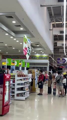 The video shows a wide shot of a busy supermarket aisle with several people standing in line at checkout counters. The aisle is well-lit with fluorescent lights and has a high ceiling with exposed ductwork. There are shelves stocked with various products on the left side of the frame. A red refrigerator is visible on the far left. A sign in the background reads "Center Souvenirs". People are mostly facing away from the camera, engaged in the process of shopping or waiting to pay. The overall impression is of a typical retail environment.