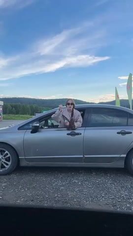 A young woman leans out of the driver's side window of a silver sedan parked on a gravel surface. The car's trunk is open. The woman is wearing sunglasses and a light-colored jacket over a darker top. Her hair is pulled back in a braid. She is holding something in her right hand and gesturing with it. The background shows a hazy sky with clouds, trees, and hills. Several tall, green and yellow flags are visible to the right of the car. The overall lighting suggests it is daytime, likely late afternoon or early evening.