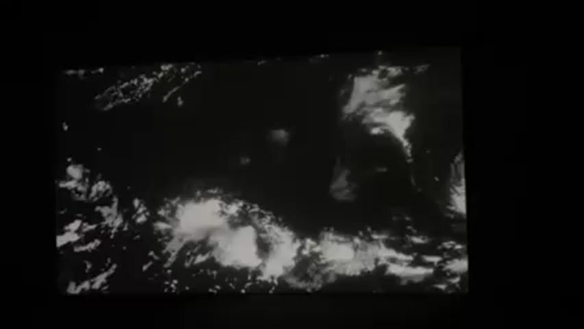 The video shows a black and white image of clouds, likely taken from space, displayed on a screen in a dark room.