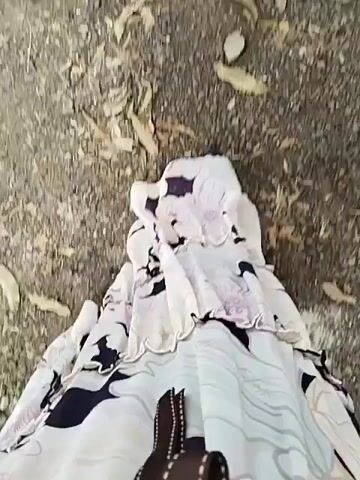 A person walks on a dirt path, their legs and a portion of their floral-patterned skirt visible. The camera is positioned to look down at the person's feet, showing their movement across the uneven ground. Dried leaves are scattered on the path.