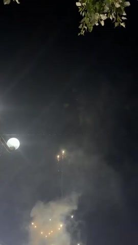 This is a vertical video of a night sky with some foliage visible at the top. A bright light, possibly a street lamp, is on the left side of the frame. Smoke and several small, bright explosions, possibly fireworks, are visible in the lower part of the sky. A string of small lights is stretched horizontally across the middle of the frame.