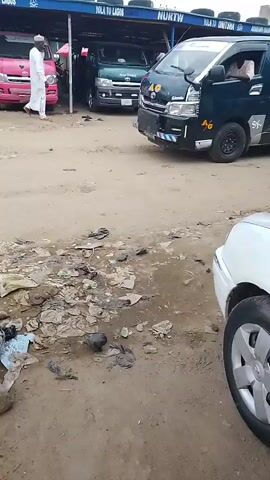 A dark green van with silver accents and the letters "AG" in yellow is parked on a dusty, debris-strewn street. The van has tinted windows and a white stripe above them. Text on the side of the van reads "NURTW" and "OLA TO LAGOS" multiple times. In the background, a pink vehicle is visible, and a man in a white shirt and blue trousers walks away from it. Another man in a white shirt and blue trousers and a white hat stands near the pink vehicle. The ground is littered with trash, including plastic bags, wrappers, and what appears to be a dead bird. A green and white striped pole stands in the foreground.