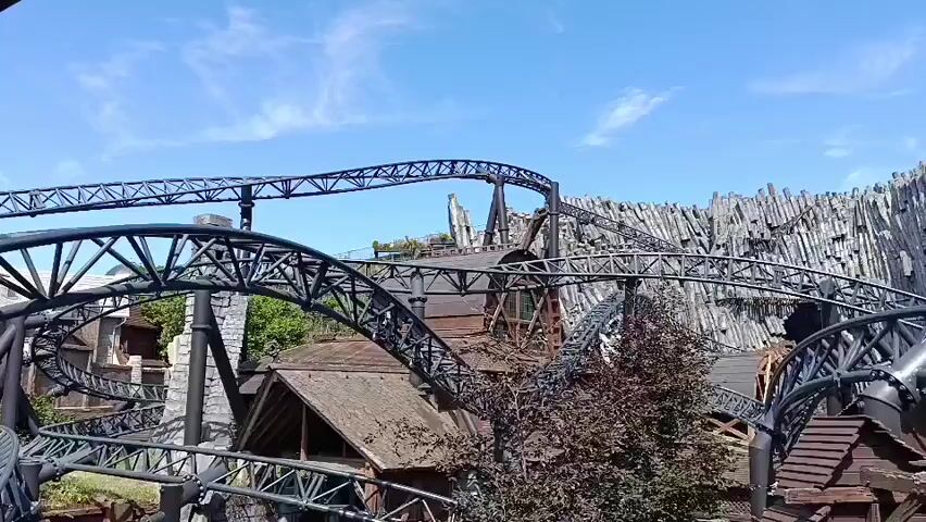 The video shows a roller coaster track winding through a themed area. The track is black and has a metal support structure. The themed area appears to be designed to resemble a village or settlement with wooden buildings and stone elements. In the background, there is a large rock formation or structure with a jagged, mountainous appearance. The sky is blue with some scattered clouds. There are trees visible within the scene. The perspective suggests the video is taken from the roller coaster itself or from an elevated position nearby. No people are clearly visible in the frame.