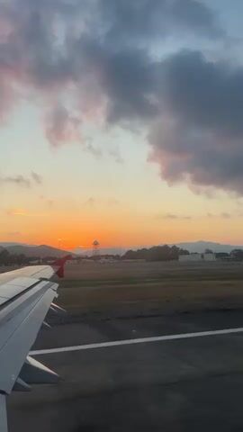 A white airplane wing with a red tip is visible on the left side, against a backdrop of an airport runway and a vibrant sunset. The aircraft appears to be moving forward on the dark grey asphalt runway, which has white markings. Beyond the runway, green grassy areas lead to distant airport buildings, including what resembles an air traffic control tower. The horizon is dominated by a bright orange and yellow sunset, with mountains silhouetted against it. Above, the sky is filled with scattered clouds, displaying hues of blue, grey, pink, and purple, illuminated by the setting sun.