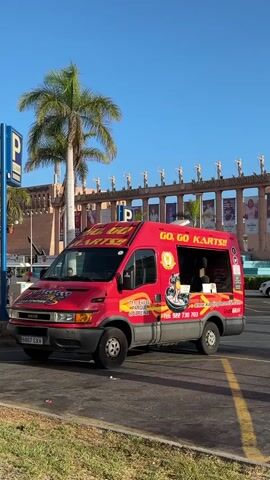 A red Iveco van with "GO, GO KARTS!!" graphics is parked on the right side of the frame. The van has a serving window open on its right side. A palm tree is on the left, and a building with statues lining its roof is in the background. The sky is clear and blue.