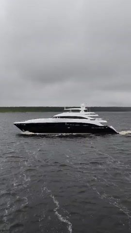 A large, modern yacht moves across choppy water. The yacht is black and white, with multiple decks and a communication tower on the top deck. It is moving from left to right in the frame, creating a wake behind it. The sky is overcast and grey. In the distance, a line of green trees can be seen. The overall scene is of a vessel traveling on a cloudy day.