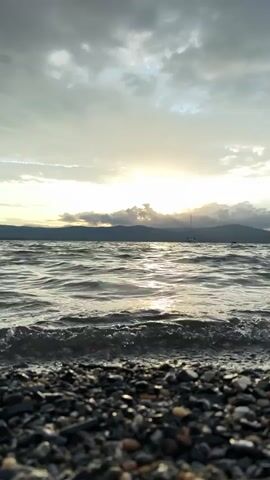 The video shows a low-angle, close-up view of a pebbled shoreline with gentle waves lapping at the shore. The sun is setting or rising, casting a warm glow over the scene and reflecting off the water. In the distance, silhouetted against the bright sky, are the shapes of mountains and a sailboat. The sky is filled with dramatic clouds, some dark and heavy, others lighter and breaking to allow the sunlight through. The overall impression is one of a serene and picturesque natural landscape.
