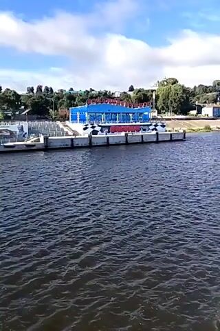 The video shows a waterfront scene with a river in the foreground and a building complex on the opposite bank. The building is modern, with a blue roof and white walls, and appears to be a public facility or a business. There are several smaller structures and what look like shipping containers in front of the main building. The riverbank is lined with concrete structures, possibly a promenade or dock. In the background, there are trees and houses, suggesting a town or city. The sky is partly cloudy, with blue patches indicating fair weather. The water is dark and rippled, with a gentle movement. There are no people or vehicles visible in the immediate frame.