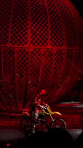 This video shows a live performance, likely in a circus or similar venue, featuring a large, red, spherical cage with a diamond-patterned exterior. Several people are present, each riding a yellow and white dirt bike. The individuals are wearing dark, protective clothing, including helmets and boots, suggesting they are performers. The floor appears to be a dark, reflective surface, possibly stage flooring, with pools of water or a wet sheen visible, reflecting the red lighting. The lighting is predominantly red, casting dramatic shadows and highlighting the performers and the cage. The performers are seen in various stages of preparation or maneuvering their motorcycles around the base of the cage, with the cage itself dominating the background. The overall atmosphere is one of anticipation and contained danger, typical of a stunt show.