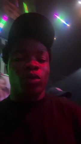 A person wearing a baseball cap in a dark environment illuminated by flashing lights. The camera is close to the person's face. In the background, other people can be seen dancing in a dimly lit club. The atmosphere appears lively and energetic.