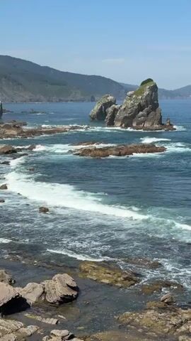 The video shows a rocky coastline with waves crashing against the shore. There are several large rock formations in the water, some of which are covered in greenery. The water is a deep blue, and the sky is clear and bright. In the background, there are rolling hills that are covered in trees. The overall scene is one of natural beauty and tranquility.