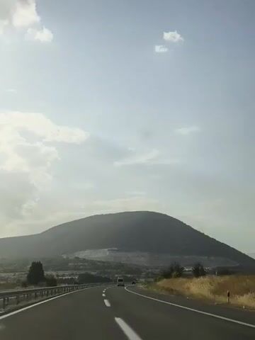 A car drives down a highway with a large, rounded hill in the background. The sky is cloudy and the sun is shining.