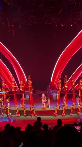 A vibrant stage performance is underway, featuring several performers in colorful costumes moving in a choreographed manner on a round stage. The stage is adorned with props resembling pillars and other decorative elements.  Overhead, illuminated red arches frame the stage.  As the performance progresses, glowing blue lotus flowers appear to float from the ceiling, creating a magical ambiance. The lighting on stage shifts between red and blue, emphasizing different aspects of the scene. An audience is present in the foreground, observing the spectacle. The camera angle is from within the audience, giving a spectator's view of the stage. The venue appears to be a theater equipped with overhead stage lighting and rigging.