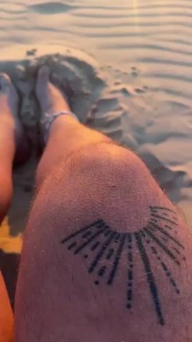 The video begins with a close-up of a person's legs and feet resting on rippled sand dunes. The person has a tattoo of a sunburst on their right thigh and a white anklet on their left ankle. The camera then pans up to reveal a serene beach scene at sunset. The sun is setting over a calm body of water, casting a warm, golden glow across the sky and reflecting on the water's surface. The sand dunes in the foreground are bathed in the soft light of the setting sun, creating a peaceful and tranquil atmosphere. The sky transitions from a clear blue at the top to vibrant oranges and yellows near the horizon. A small sailboat can be seen in the distance on the water. The overall mood is one of relaxation and natural beauty, suggesting a moment of quiet contemplation enjoyed at the end of a day. The scene evokes a sense of warmth and serenity, likely experienced on a beach in La Teste-de-Buch, France.
