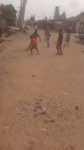 A group of young boys are playing soccer in a dirt road. The boys are wearing casual clothing. The environment appears to be a village or rural area with simple buildings visible in the background. The sky is overcast. The video shows the boys running, kicking the ball, and interacting with each other as they play.