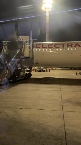 A video shows an airplane on the tarmac at night. The airplane is white with red lettering that says "ELECTRA". A jet bridge is attached to the airplane's door, and people are disembarking from the airplane via the jet bridge. The ground is asphalt, and there are lights illuminating the scene. A tall light pole with a bright light at the top is visible in the background. The video appears to be filmed from inside another building, possibly an airport terminal, as a reflection is visible on the glass.