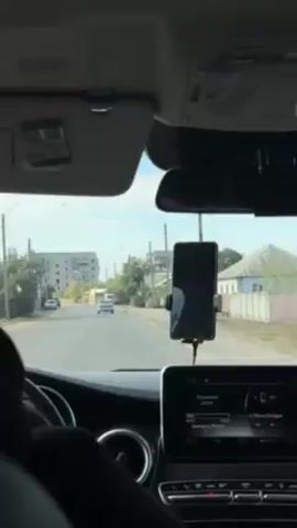 The video shows the interior of a car, specifically the driver's perspective. The car is driving down a residential street on a sunny day. Other vehicles and buildings can be seen in the distance. A smartphone is mounted on the dashboard, and the car's infotainment system displays music information. The driver's arm and part of their shoulder are visible on the left side of the frame. The video is likely taken from a smartphone, possibly for recording a drive or navigating.