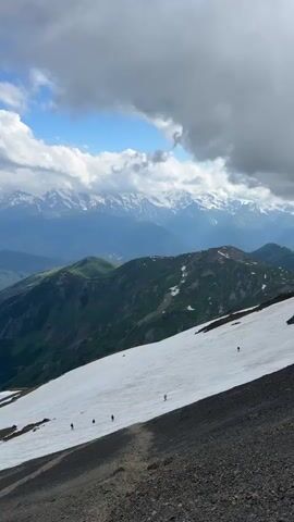 The video shows a group of people hiking up a snowy mountain slope. The sky is partly cloudy with some patches of blue. In the background, there are more mountains, some of which are covered in snow. The hikers are spread out along the slope, and some appear to be using trekking poles. The ground in the foreground is rocky and barren. The overall impression is one of a challenging but rewarding outdoor adventure.