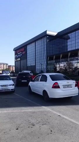 The video captures a modern building with large glass windows and an arched entrance. A white car is parked in front of the building. The scene appears to be an outdoor area, possibly a shopping center or a commercial complex, with some greenery and an orange cone visible. The weather seems sunny with a clear blue sky. The text "KO'K SAROY SAVDO MAJMOSI" is visible above the entrance, indicating the name of the establishment. There are a few people walking near the entrance.