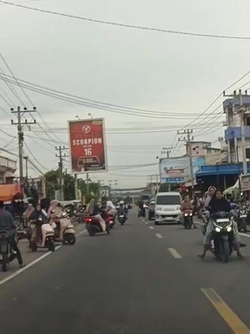 The video shows a busy street scene with multiple lanes of traffic, predominantly motorcycles and some cars. The weather appears overcast. On the left side of the road, there are several parked motorcycles and scooters, with some people standing around them. A market stall or shop with goods is visible behind them. A large billboard advertising "RED SCORPION 16" is prominently displayed overhead. On the right side of the road, buildings line the street, and there are more motorcycles and a white van in motion. The road itself is paved and has yellow lane markings. Power lines and utility poles are visible throughout the scene, indicating an urban or semi-urban environment. The overall impression is one of a lively and active street, likely in Southeast Asia, given the prevalence of scooters and the attire of some individuals.