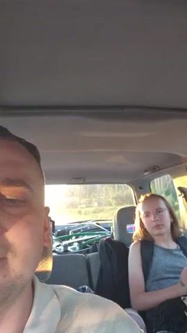 The video is filmed from inside a vehicle. In the foreground, on the left, a man's face is visible, partially illuminated by sunlight. In the background, on the right, a young person wearing glasses and a gray tank top is seated. The person is looking towards the camera and appears to be speaking. Behind the seats, the interior of the vehicle is visible, including car seats and luggage. Through the windows, a road and trees are visible, with sunlight streaming in. The overall lighting suggests it is either sunrise or sunset.