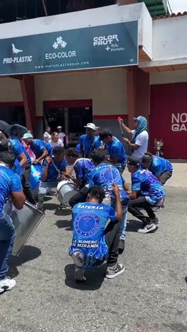 A group of people wearing blue shirts and jeans are playing drums in the street. Some members are kneeling, while others are standing. There is a crowd of people, including children, walking behind them. Most people in the crowd are waving blue and white balloons. The event seems to be a parade or march of some kind. There are several stores and business signs visible in the background. The weather is sunny and bright.