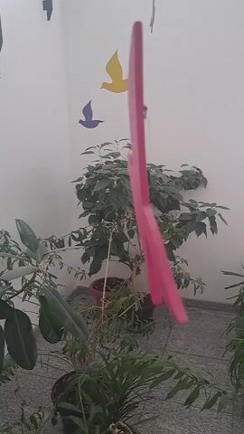 A pink bird-shaped mobile hangs in front of a white wall and several potted plants. Two smaller bird cutouts, one blue and one yellow, are adhered to the wall behind the mobile. The mobile appears to be made of flat, rigid material and is suspended by a thin string. It is positioned in the center of the frame and takes up a significant portion of the image. The plants are lush and green, and the floor appears to be tiled. The lighting is bright, suggesting it is daytime and possibly outdoors or in a well-lit indoor space.