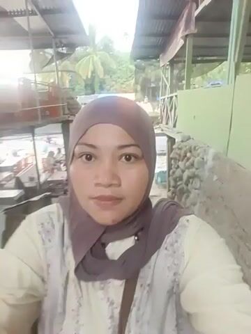 A woman wearing a hijab and a white patterned shirt is standing in front of a camera, likely recording a selfie video. The background suggests an outdoor setting with some structures that appear to be stalls or small buildings with tin roofs. There are tables and chairs visible, some with people seated at them, indicating a public area, possibly a market or a food court. The ground is uneven and appears to be made of gravel or dirt. In the distance, a body of water and trees can be seen, suggesting a natural environment. The lighting is somewhat diffused, indicating it might be daytime. The woman is looking directly at the camera and appears to be speaking or interacting with it.