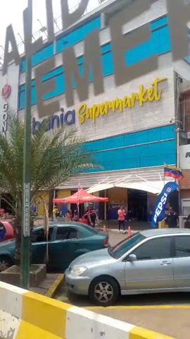 The camera is in a moving vehicle, looking at a busy commercial street. The main feature is a large building with the names "GONAVI" and "Bahia Super" on it, indicating a shopping complex. Several motorcycles are parked in front. People are walking by and some are waiting at a bus stop. The bus stop has an advertisement featuring two men. A palm tree in a concrete pot is seen at the beginning of the clip.