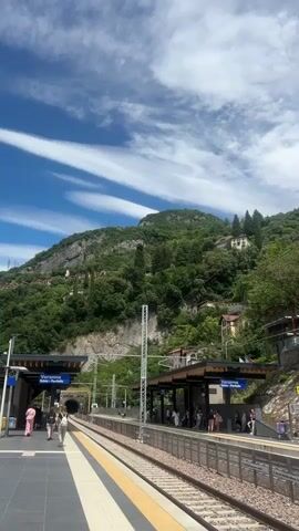 The video shows a train station with tracks and platforms. There are several people standing on the platforms, some appearing to be waiting for a train. A tunnel is visible in the distance. The station is situated at the base of a steep, green hillside dotted with houses and trees. The sky is blue with white, wispy clouds. The overall impression is a scenic and tranquil setting.