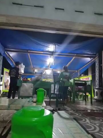 The video shows an outdoor seating area at night, likely a food stall or small restaurant. A blue tarp covers the structure, supported by wooden beams. Several light bulbs illuminate the area. Two individuals are visible, one in a black shirt and the other in a green shirt. They appear to be working behind a counter or food preparation area. Tables and green chairs are set up in front of the counter, some covered with patterned mats. A large green thermos is prominently placed on a table in the foreground. The ground is tiled. The overall impression is of a humble, casual dining establishment.