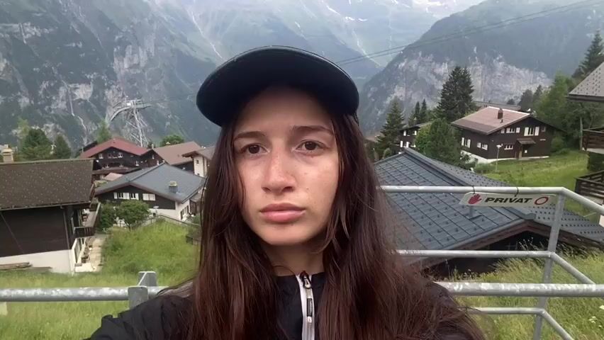 A young woman with long, dark hair and a black baseball cap stands close to the camera, facing forward. She wears a black zip-up jacket. Behind her, a village of wooden chalets with red roofs is nestled on a green mountainside. In the background, snow-capped mountains rise dramatically under a cloudy sky. The image is a selfie, with the woman's arm and hand visible at the bottom left, holding the camera. A sign with the word "PRIVAT" is visible on a railing to the right.