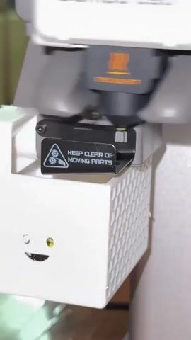 This is a close-up, vertical, eye-level shot of a 3D printer's print head and the upper portion of a white accessory attached to it. The print head, labeled "BAMBOO LAB" and "CAUTION HOT," is dark gray and black, with an orange symbol indicating heat. Below the print head is a black rectangular component with a yellow wire extending from it. A black sticker with a warning symbol of gears and the text "KEEP CLEAR OF MOVING PARTS" is affixed to this component. The white accessory below has a patterned texture on its right side and a raised, cartoon-like emoji of a pile of excrement on its front. The emoji has two eyes and a smiling mouth, with one eye appearing yellow. The lighting is bright, and the background is a blurred light color. The video shows subtle, rapid movements of the print head and the yellow wire.