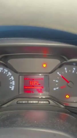 A car dashboard is visible, with a red digital display showing "105". A tire pressure warning light is illuminated. The view then shifts to a road seen through the windshield, with streetlights and fields lining the road. The sky is a twilight blue.