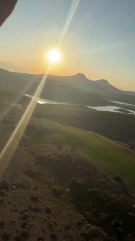 The vertical video captures an early morning hot air balloon flight over a scenic landscape in Yeşilhisar, Turkey. The video opens with a bright sunrise, casting long rays of light over a mountainous terrain. A large body of water, likely a lake or reservoir, is visible between the rolling hills and mountains in the distance. Below, a patchwork of lush green fields and drier, brush-covered land stretches across the foreground. As the camera pans to the right, two hot air balloons come into view. One prominent yellow and red balloon ascends gracefully against the clear blue sky, while another can be seen further in the distance. The weather is clear and sunny, indicating an ideal day for flying. The scene is serene and peaceful, highlighting the beauty of the natural landscape and the unique experience of a hot air balloon ride at dawn. No visible text is present.