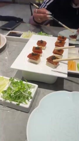 The video shows a close-up of a table in what appears to be a restaurant. There are several dishes of food, including what looks like Peking duck, served on white rectangular plates. Two sets of chopsticks are holding a piece of Peking duck, with one set about to place it into a soy sauce dish. Other small dishes contain sliced scallions and microgreens. In the background, a bamboo steamer basket filled with food and a bowl of grapes are visible. The table is made of a grey, marbled material. A person's hand with manicured nails can be seen in the upper right corner of the frame, holding chopsticks. The overall lighting suggests an indoor setting, possibly a dimly lit restaurant.