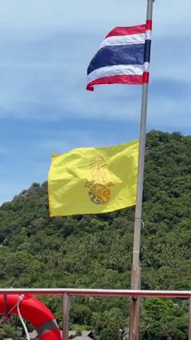 The video shows a close-up of a flagpole with two flags. The upper flag is the national flag of Thailand, with red, white, and blue stripes. The lower flag is yellow and features a royal emblem, likely the Royal Cypher of the King of Thailand. Both flags are waving in the wind. Behind the flagpole, a lush, green, tree-covered hillside is visible under a bright blue sky with scattered clouds. In the foreground, part of a boat railing and a red life preserver are visible. The video is taken from the perspective of someone on the boat.