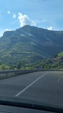 The video shows a view from inside a car, looking out at a winding road and a mountainous landscape. The road is asphalt with a white line marking the lanes. A guardrail runs along the side of the road. The mountains are covered in green vegetation, and the sky is blue with scattered white clouds. Sunlight is streaming down, creating streaks of light across the mountains. The video appears to be filmed while the car is in motion, as the landscape is slightly blurred. There are no people or other objects visible besides the road, guardrail, mountains, and sky.